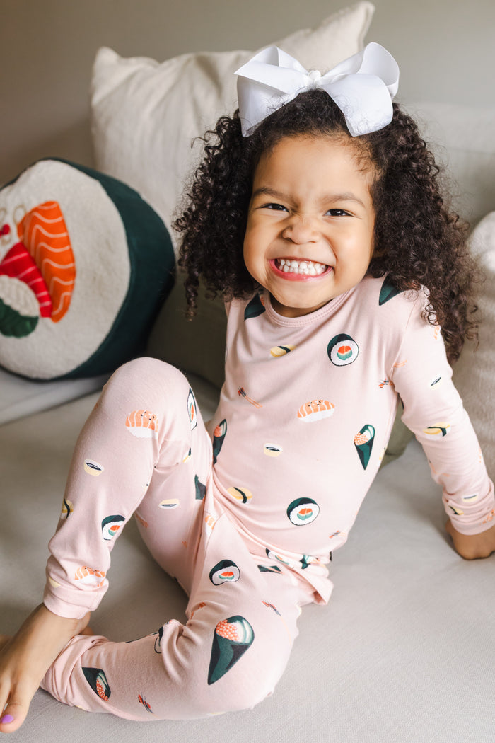Child smiling sitting on a white couch  with one leg up wearing blush sushi pajama set with a white bow