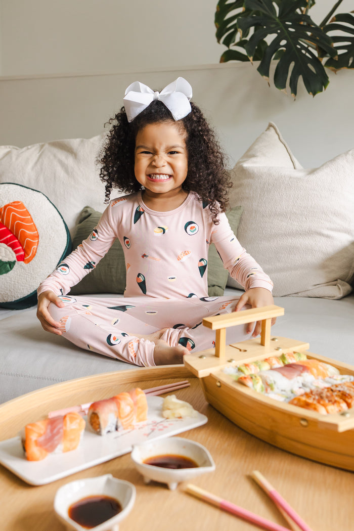 Child sitting crisscross on a white couch  wearing blush sushi pajama set and white bow with sushi on a table in front of her