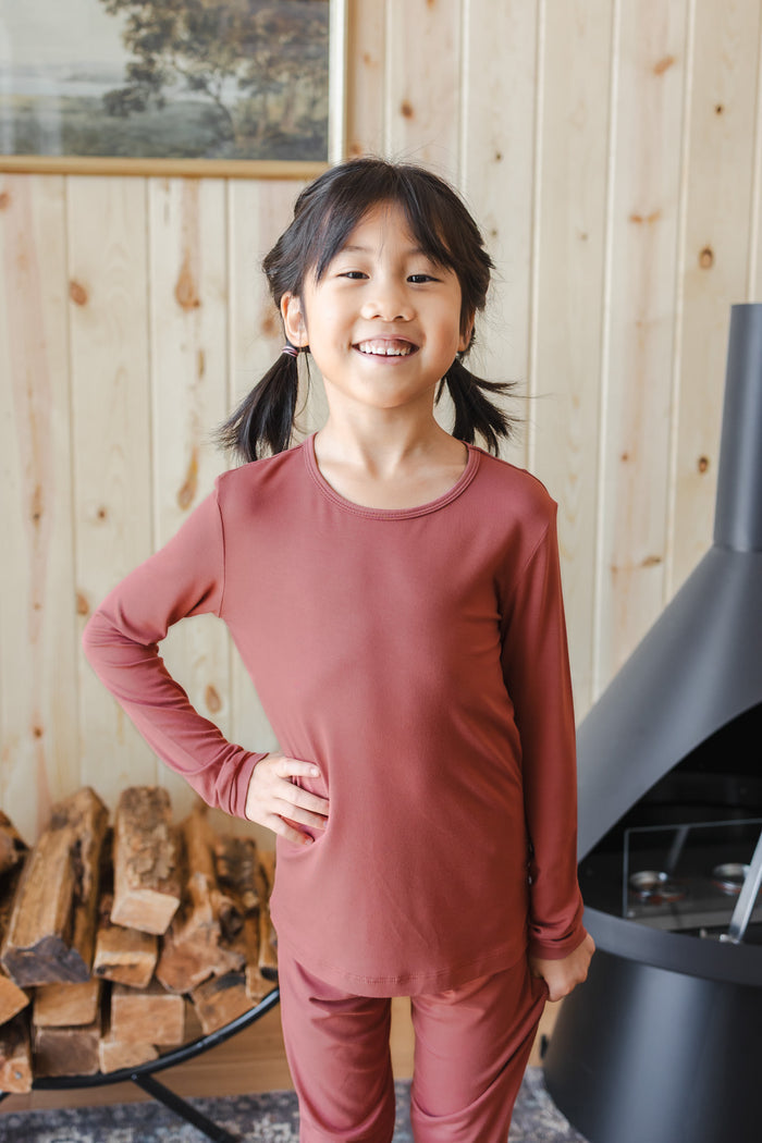 A young girl with dark hair in pigtails stands smiling in a cozy room with light wood-paneled walls, wearing a matching dusty terracotta long-sleeve top and pants pajama set.