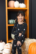 Girl smiling in black Long Sleeve Pajamas in Halloween Friends print sitting with pumpkin decorations