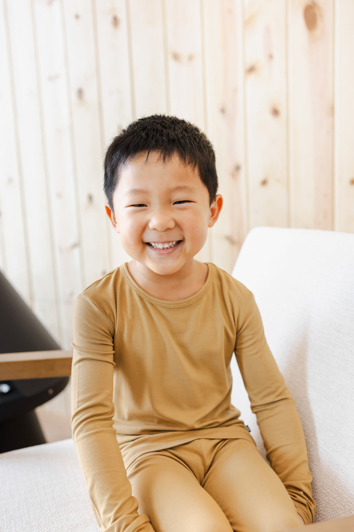 A young boy smiling while wearing a matching mustard yellow long-sleeve pajama set, sitting on a white sofa against a light wood-paneled wall.