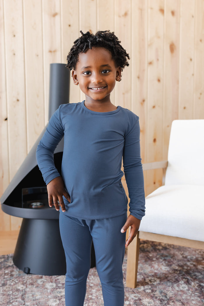 A young child wearing a matching slate blue long-sleeve pajama set, standing in a modern living room with a wood-paneled wall, a black fireplace, and a white chair.