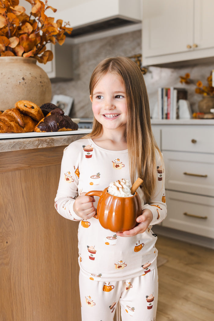 Girl smiling in kitchen wearing Long Sleeve Pajamas with Pumpkin Spice Latte print holding a spice latte cup.