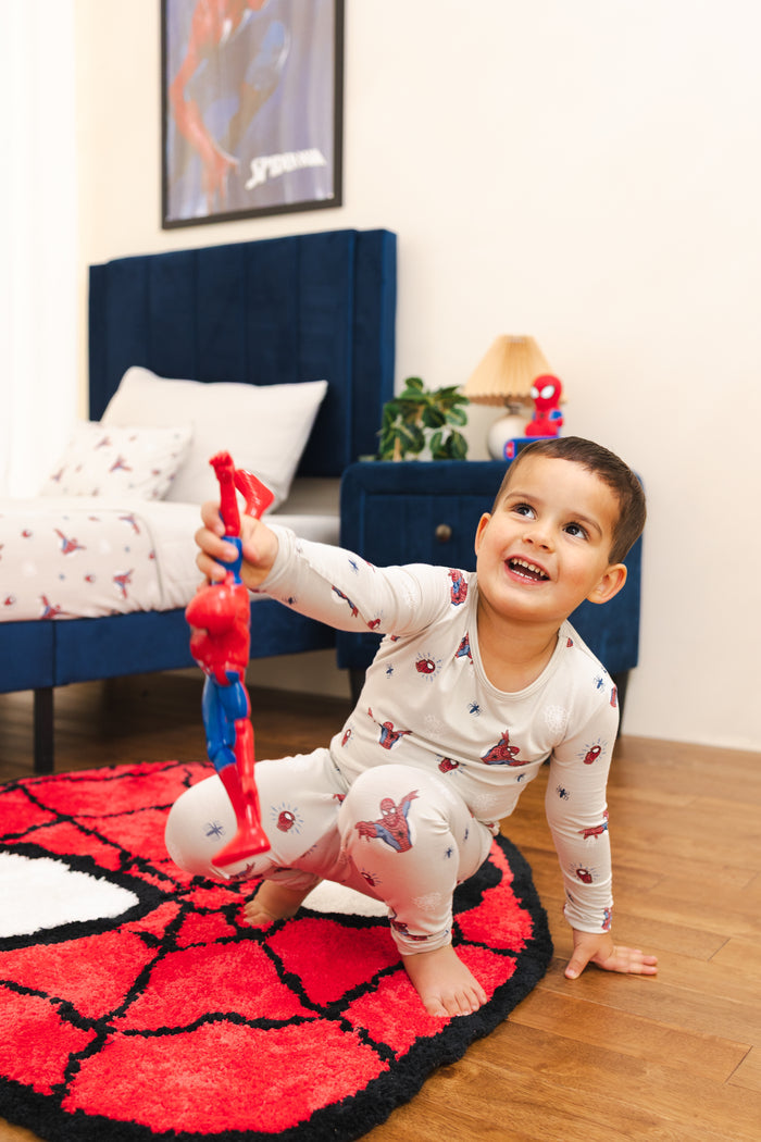 A young boy wearing a Spider-Man themed pajama set crawls on a red and black Spider-Man themed rug in a bedroom, holding a Spider-Man toy.