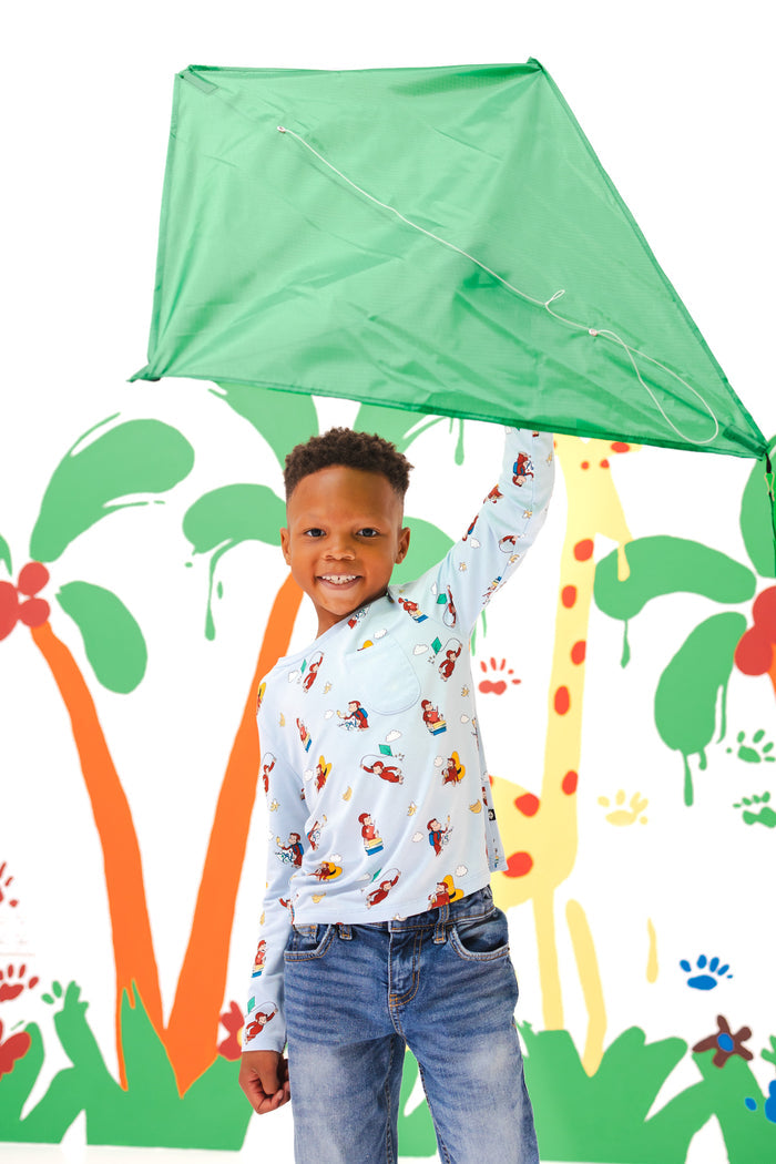 Boy holding a kite standing in front of a colorful background wearing Long Sleeve Toddler Crew Neck Tee in Curious George print over a blue jeans