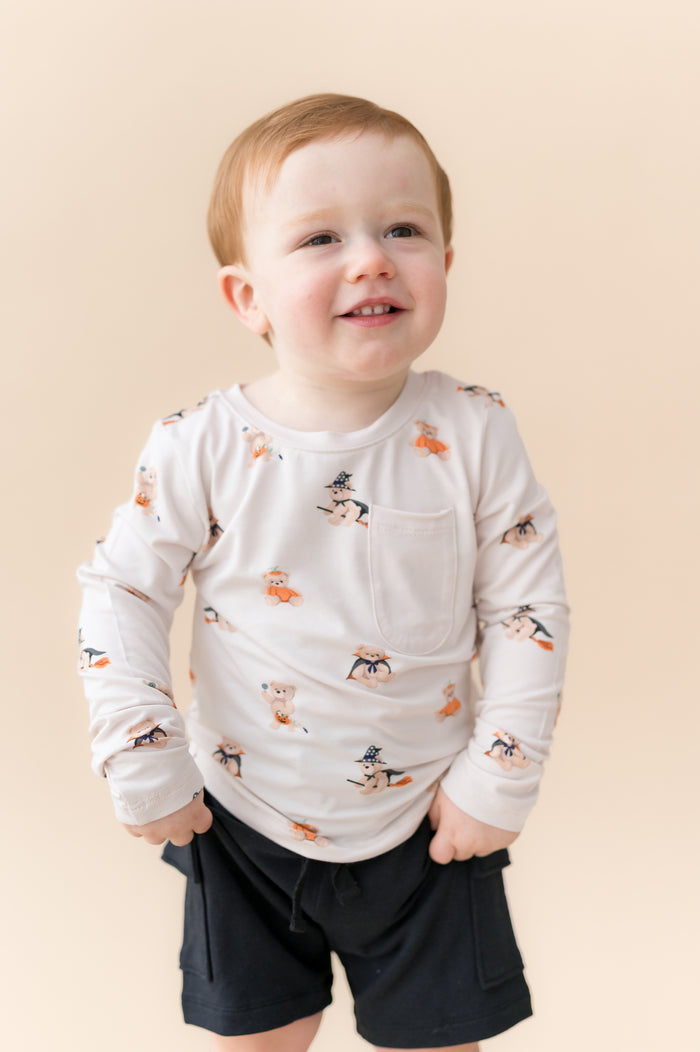 Boy posing in Long Sleeve Toddler Crew Neck Tee in Halloween Teddy print over black shorts on a beige background