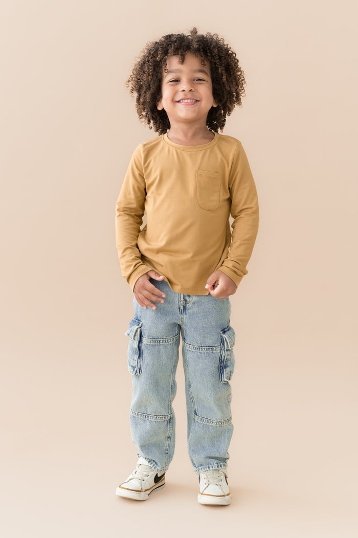 Boy smiling in Long Sleeve Toddler Crew Neck Tee in Ochre over blue jeans on a beige background