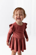 A toddler girl with light brown hair wearing a dusty terracotta long-sleeve dress with ruffled shoulder details, standing against a bright white background.
