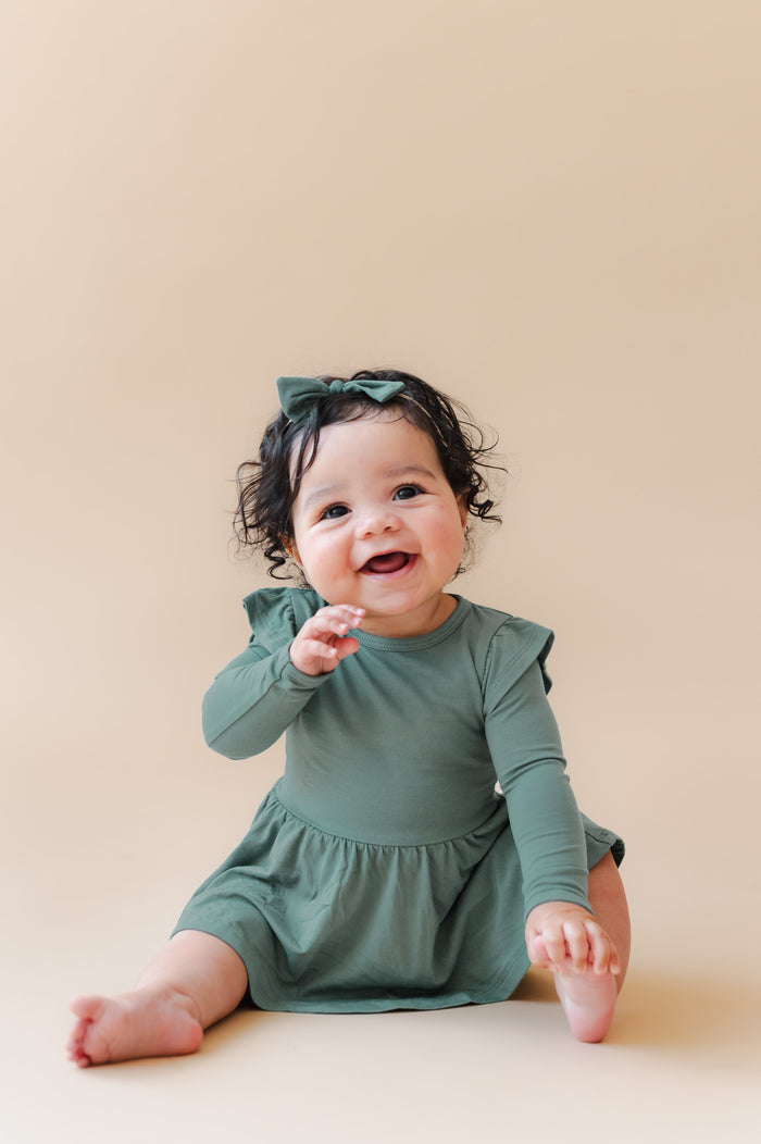 A smiling baby girl sitting on a beige studio floor, wearing a sage green long-sleeve bodysuit dress with ruffled shoulders and a matching green bow headband.