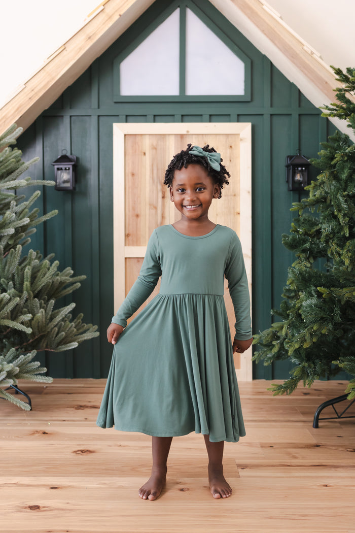 Girl wearing Long Sleeve Twirl Dress in Pine standing in front of a beige door