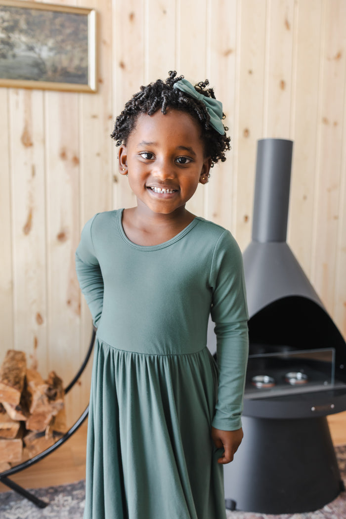 Girl smiling in Long Sleeve Twirl Dress in Pine standing near chimney