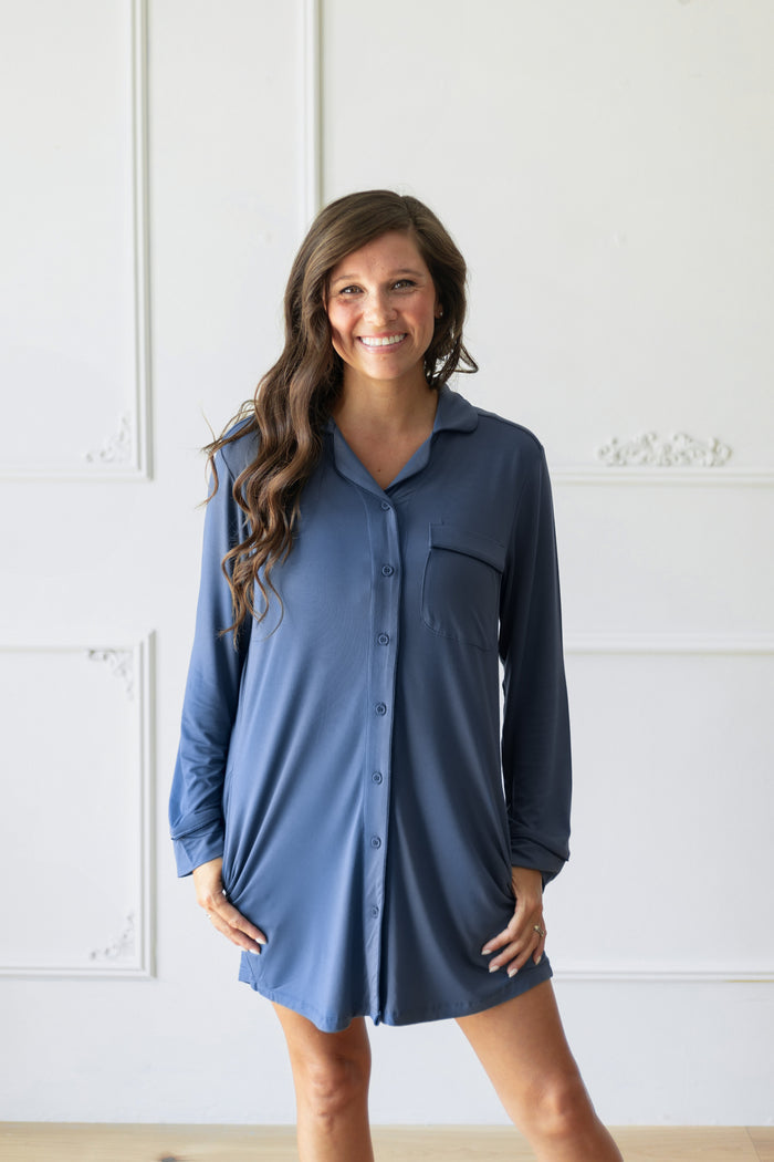 A woman wearing a slate blue long-sleeve button-down night shirt dress, standing against a white paneled wall.