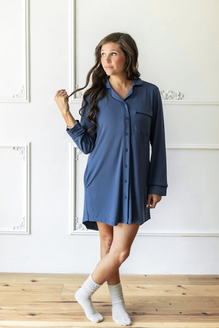 A woman wearing a slate blue long-sleeve button-down shirt dress, styled with white socks, standing in a room with white paneled walls and light wood floors.