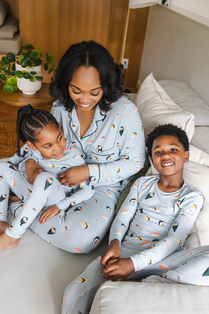 A woman and two children wearing matching light blue pajamas with a colorful sushi print, sitting together on a light-colored sofa in a cozy living room setting.
