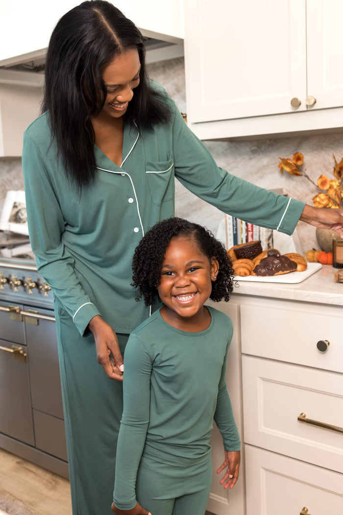 A woman and a young girl wearing matching sage green long-sleeve pajamas with white piping, standing in a bright kitchen.