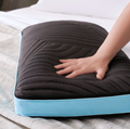 A hand presses down on a Malouf Ice Cloud™ Pillow with a light blue border on a bed with white sheets. The pillow, from the brand Malouf, is made of plush memory foam and has a textured surface with wavy lines. The light blue piping contrasts with the black fabric, and the mattress and part of a white pillow are visible in the background.