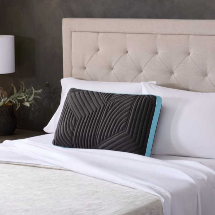 A neatly made bed featuring a tufted beige upholstered headboard. On the white pillows is a prominently placed Malouf Ice Cloud™ Pillow from Malouf, showcasing a geometric pattern and an aqua blue edge. The bed, enhanced with plush memory foam, has a white duvet and sheets. A white nightstand with a lamp and potted plant sits to the left.