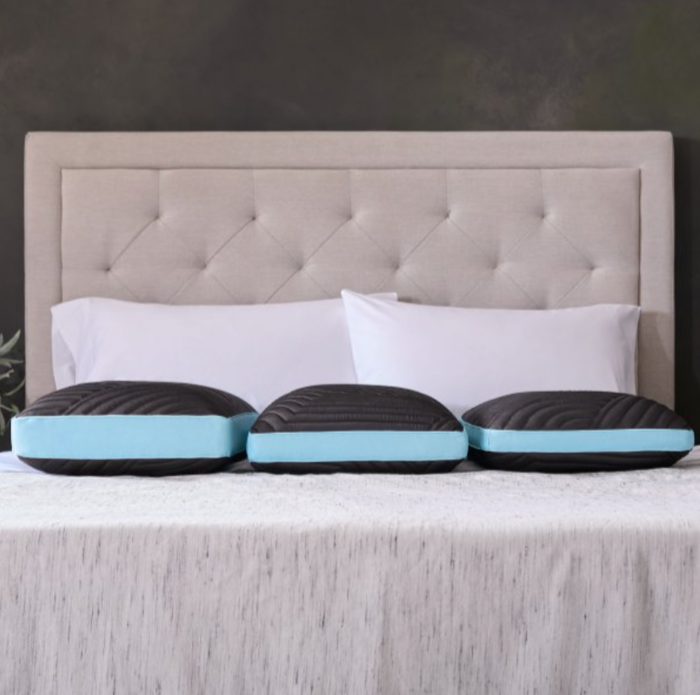 A beige, tufted headboard stands against a dark wall, with two white pillows placed in front of it. On the light gray bedspread below, three identical Malouf Ice Cloud™ Pillows by Malouf, featuring black and blue memory foam with cooling gel technology, are neatly aligned, enhancing the modern and cozy bedroom ambiance.
