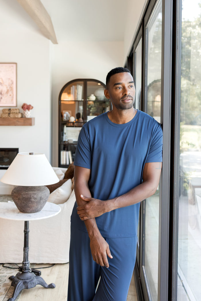 Model posing in Men's Crew Neck Tee in Oxford over Men's Lounge Pants i oxford standing against a window in a living room
