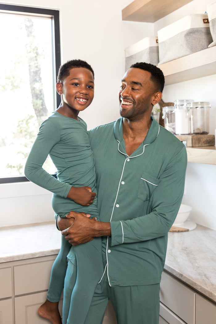 A man and a young boy wearing matching sage green pajamas with white piping, standing together in a bright kitchen setting.