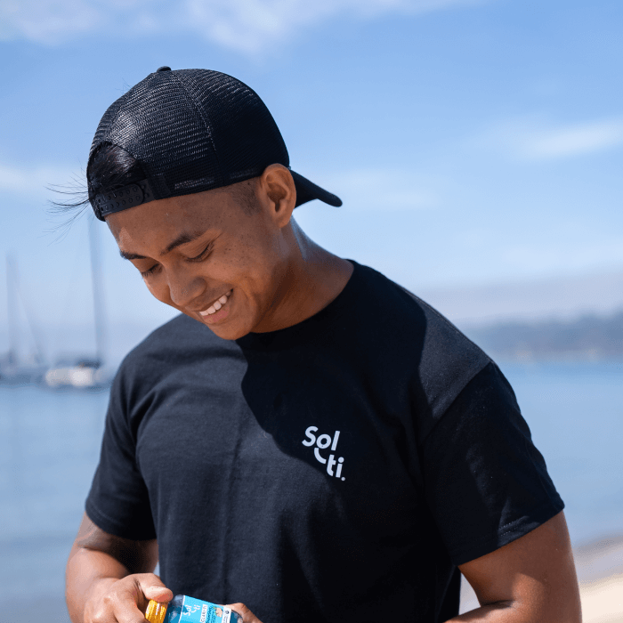 A man wearing a black Sol-ti organic cotton t-shirt and a black cap, holding a Sol-ti SuperShot shown against a light blue background.