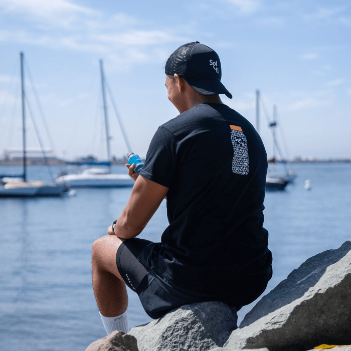 A man wearing a black Sol-ti organic cotton t-shirt and a black cap sitting in a rock on the beach with boats nearby, holding a Sol-ti SuperShot