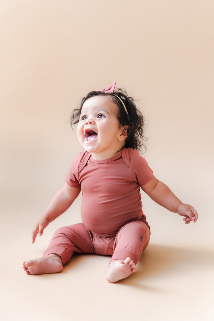 Baby wearing Pant in Ember over similar color bodysuit and bow headband on a beige background