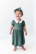A toddler girl wearing a sage green dress with a white Peter Pan collar, matching green bow headband, and standing barefoot against a white studio background.
