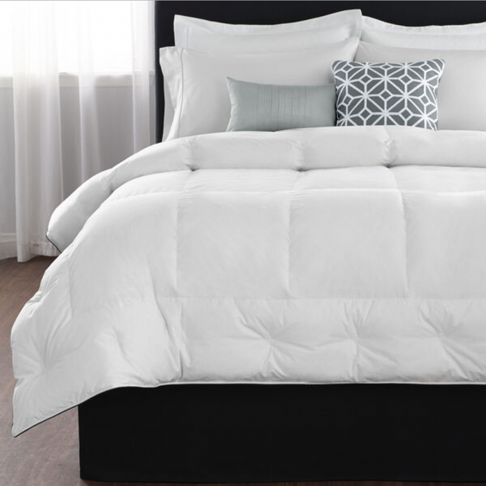 A neatly made bed with a Restful Nights® Euro Box Down Alternative Comforter and several pillows, including one with a geometric pattern in grey and white, and a smaller plain grey pillow. The headboard is upholstered in a dark material, and sheer white curtains are drawn to either side of a large window in the background.
