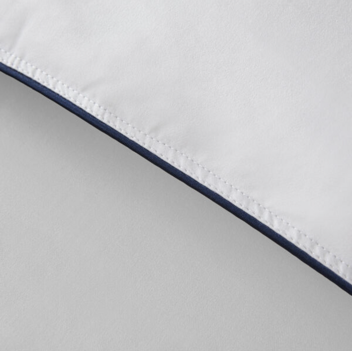 Close-up of a corner of a white pillow with navy blue piping along the edge. The stitching is visible along the seam, reminiscent of the detailed craftsmanship found in the Restful Nights® Euro Box Down Alternative Comforter. The fabric appears smooth and clean, with a subtle shine in the lighting. The background is a plain, light gray surface.