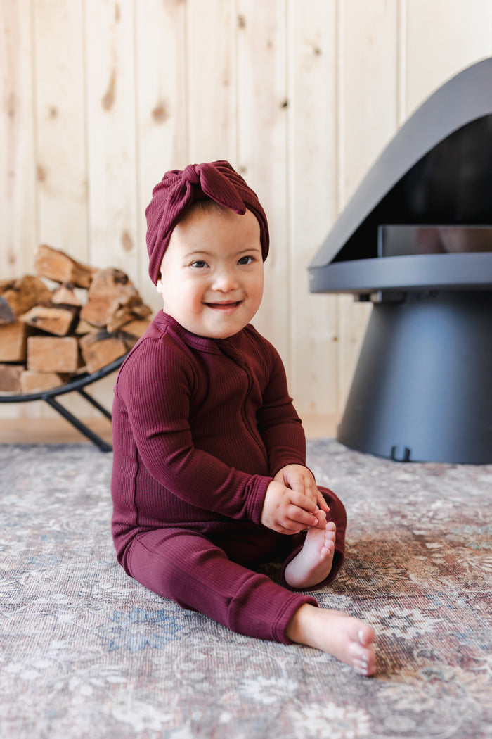 Toddler wearing Ribbed Zipper Romper  and Ribbed Headwrap in Burgundy sitting on carpet
