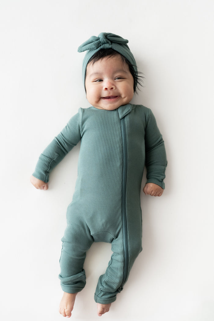 Baby wearing Ribbed Headwrap in Pine with zippered romper on a beige background
