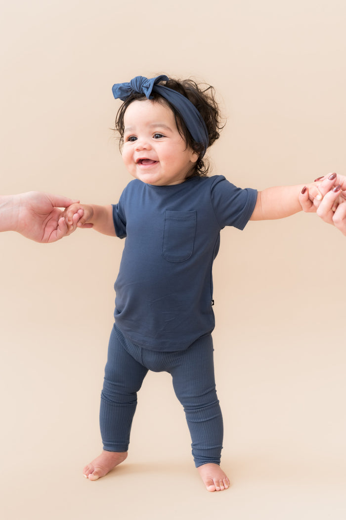 Baby wearing Ribbed Leggings and knotted bow head band in Oxford holding a hand on a beige background