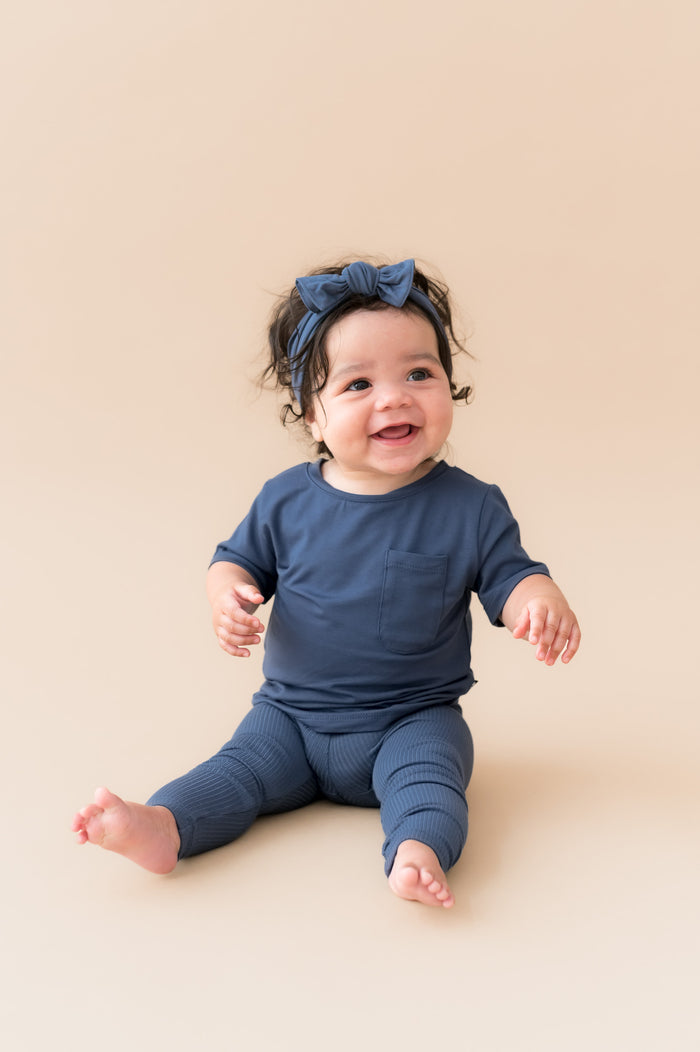 Baby smiling in Ribbed Leggings and knotted bow headband in Oxford on a beige background