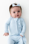 Infant girl wearing the Ribbed Ruffle Zipper Footie in Breeze with matching tiny nylon bow
