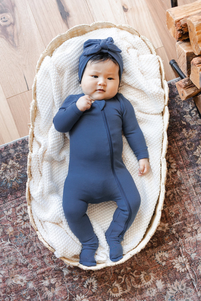Baby wearing Ribbed Zipper Footie and Ribbed Headwrap in Oxford lying in a basket