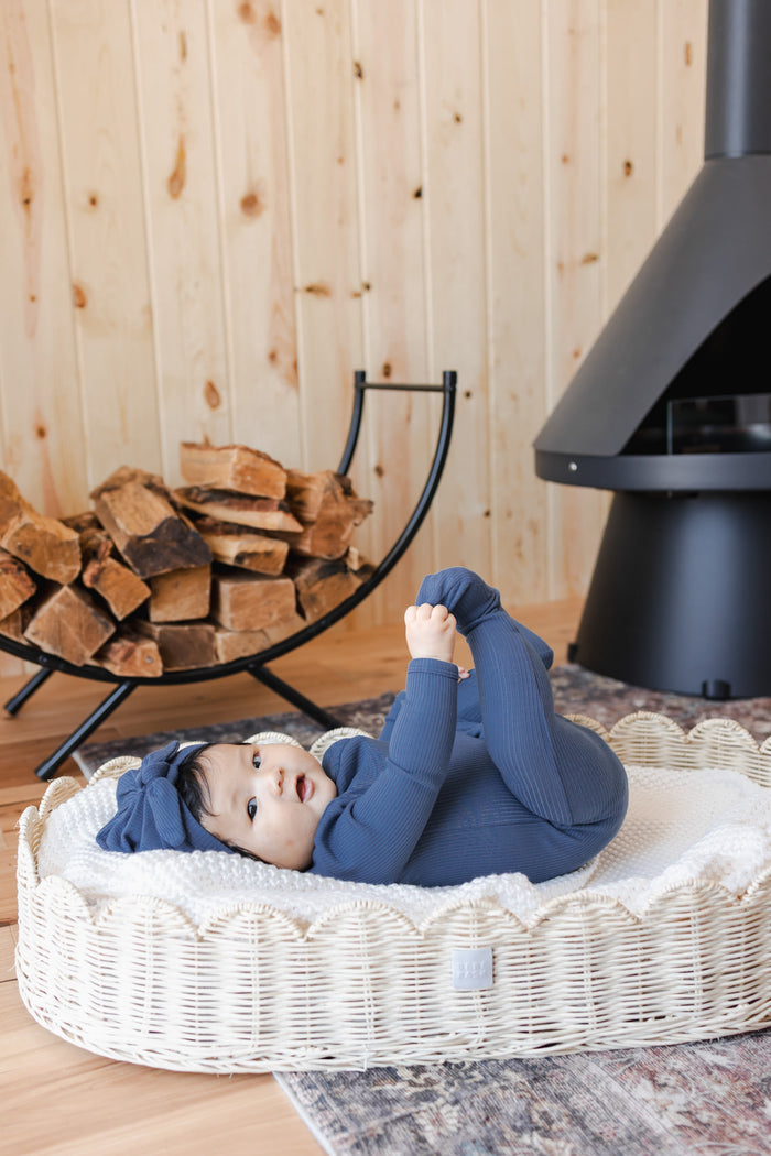 Baby holding her feet wearing Ribbed Zipper Footie and Ribbed Headwrap in Oxford lying on a basket