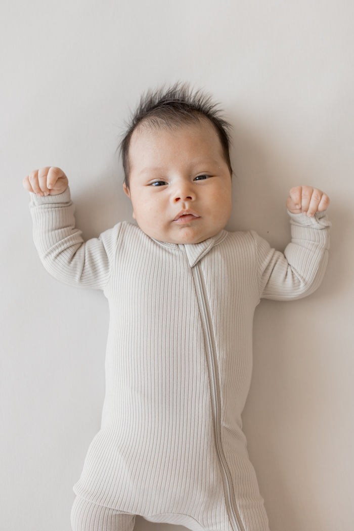Close up of Baby wearing Ribbed Zipper Romper in Oat color on a beige background