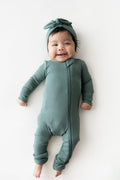 A smiling baby lying on their back wearing a ribbed sage green footless one-piece with a matching knotted headband, set against a plain white background.