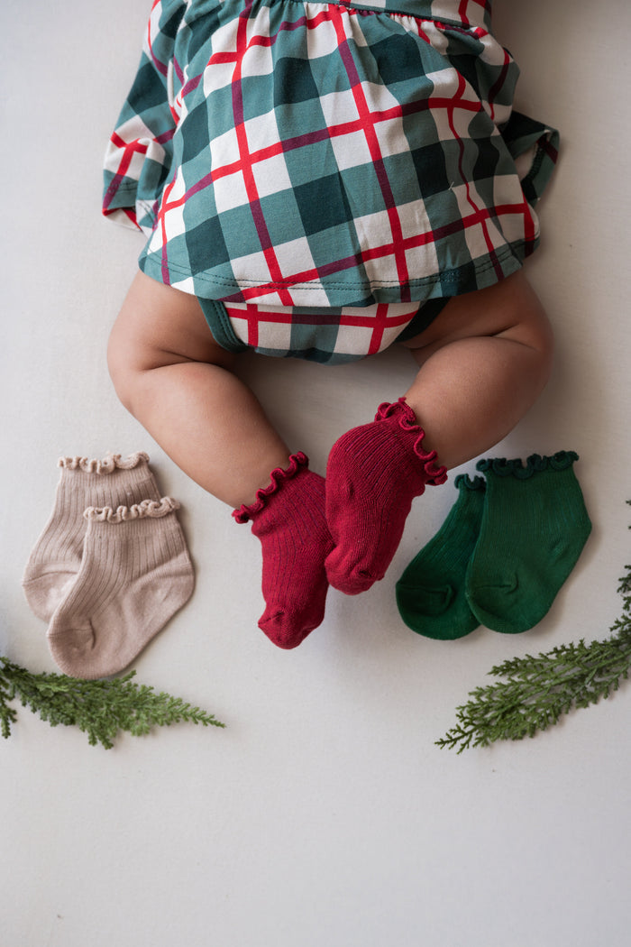A baby lying on a white surface wearing a green, white, and red plaid outfit, with three pairs of ruffled socks displayed around them: one beige pair, one red pair, and one green pair. Green pine branches are placed in the corners.