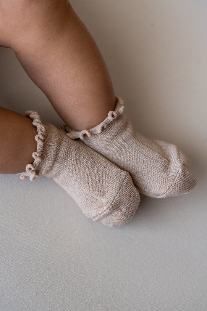 A close-up of a baby's feet wearing light beige ribbed knit socks with ruffled trim at the top, resting on a neutral light gray surface.