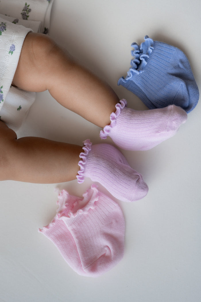 Baby wearing light purple ruffle socks with 2 other socks laying next to them in a periwinkle and light pink color