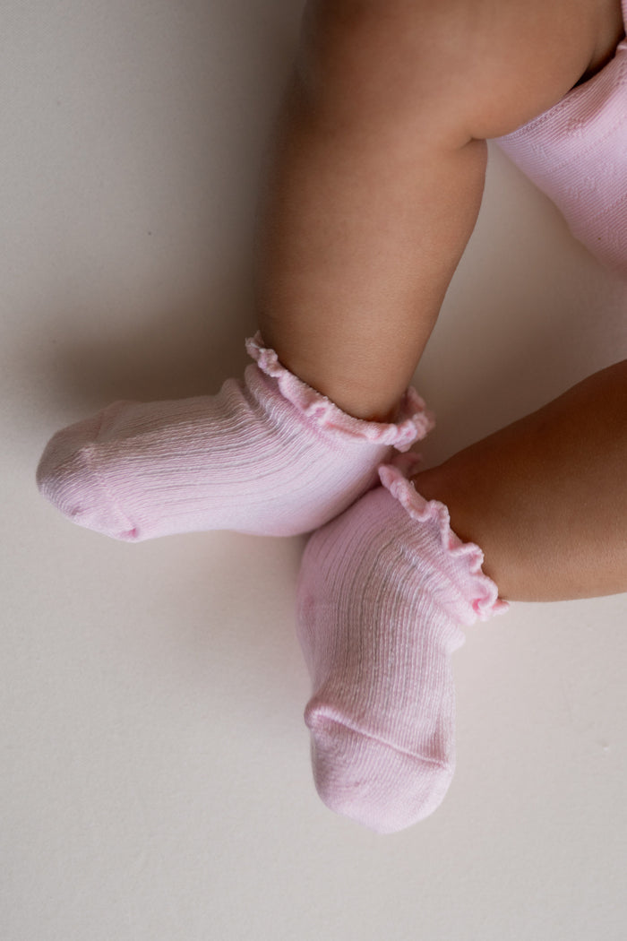 A close-up of a baby's legs wearing light pink ribbed ankle socks with ruffled trim, resting on a light beige surface.
