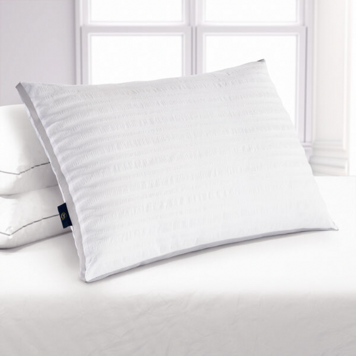 The Serta Dream Loft Pillow by Keeco, featuring a subtle vertical stripe pattern on its super soft white seersucker fabric, is centrally placed on a neatly made bed with white linens. Another pillow is partially visible behind it. The background showcases bright windows with a minimalist design, allowing natural light to illuminate the scene.