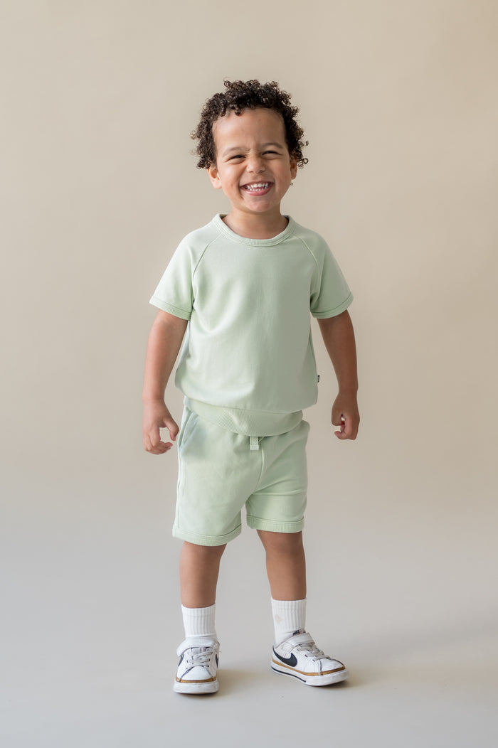 Smiling boy wearing the Short Sleeve Jogger Set in Basil in front of a light beige background