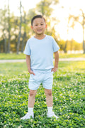 Young boy standing in a grass field wearing the Short Sleeve Jogger Set in Breeze with his hands in the pockets of the shorts