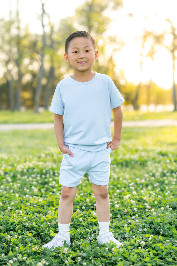 Young boy standing in a grass field wearing the Short Sleeve Jogger Set in Breeze with his hands in the pockets of the shorts