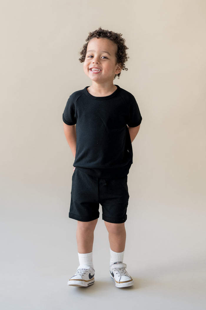 Young boy smiling with his hands behind his back wearing the Short Sleeve Jogger Set in Midnight