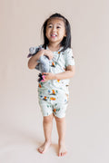 Girl smiling in Short Sleeve Pajamas in Winnie the Pooh Hundred Acre Wood print holding a stuff toy on a cream background