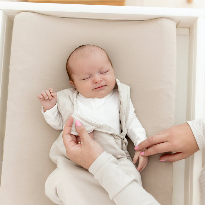 Sleeping infant laying on a changing pad in Oat wearing Kyte Baby Sleep Bag in Oat 1.0 while it is being unzipped